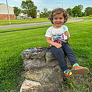 Timmy is registered to the contest to win money with this photo: boy, child, clothing, face, footwear, grass, head, male, nature, outdoors, pants, park, person, photography, plant, portrait, rock, shoe, sitting, tree