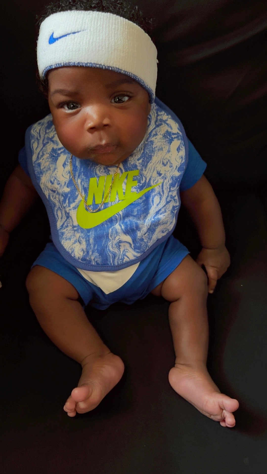 Andre Henry Jr is registered to the contest to win money with this photo: baby, infant, headband, nike, bib, blue_clothing, sitting, barefoot, dark_background, cute, child, portrait, person, indoors, expression, skin, foot, hand, face, young