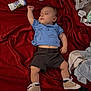 baby, sleeping, blue_shirt, shorts, beige_sneakers, red_blanket, water_bottle, socks, remote_control, blanket, toddler, clothing, footwear, indoor, resting, infant, casual, sleep, child, relaxing