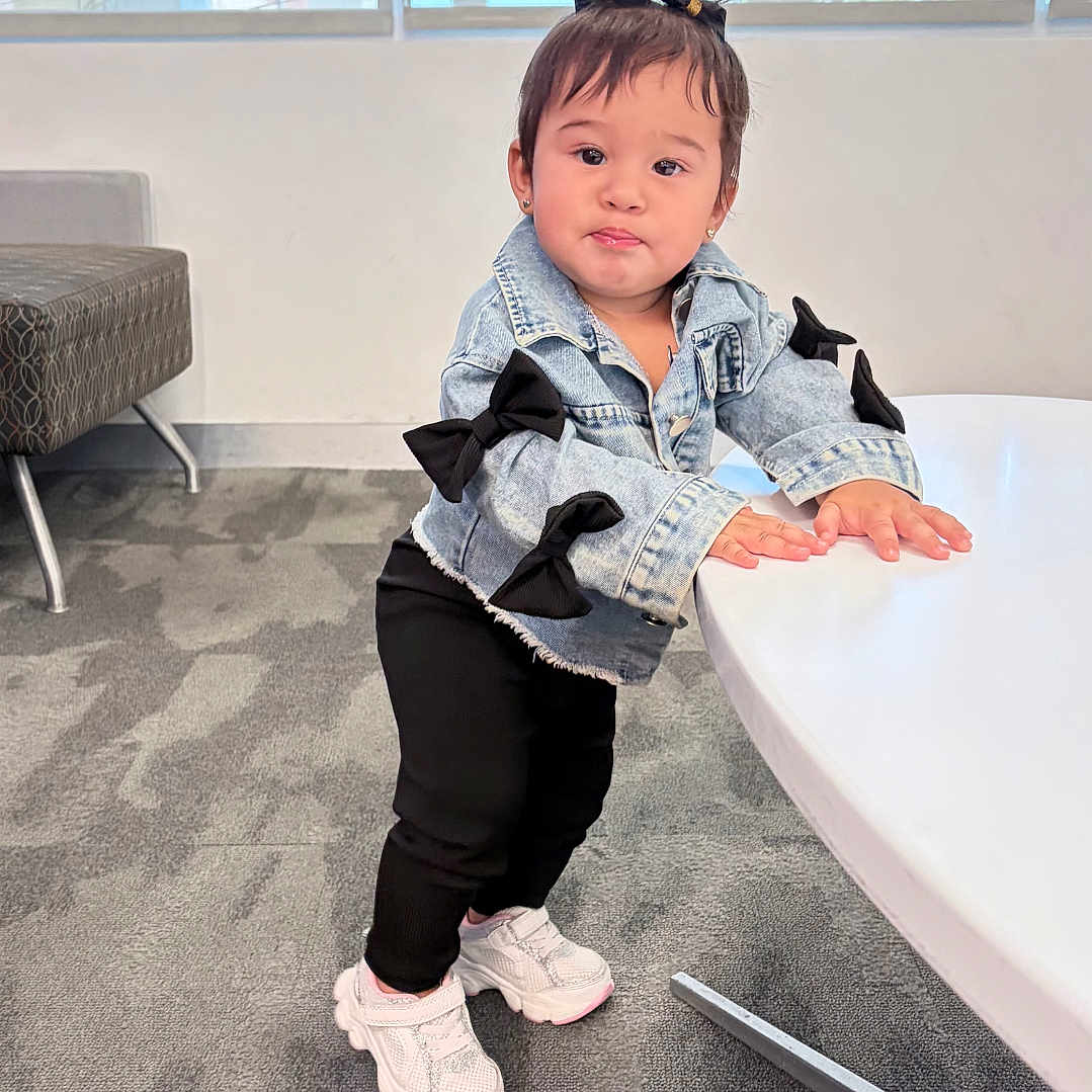 Elif joined the competition — help win amazing prizes! toddler, child, denim_jacket, black_bows, standing, white_table, carpet, indoor, window, light, curious, sneakers, black_pants, hair_bow, cute, playful, person, young_child, casual_wear, modern_interior