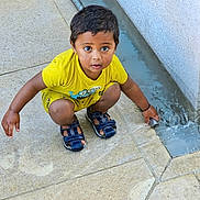 Riyansh a rejoint le concours — aidez-le/la à gagner de superbes lots ! child, boy, yellow_clothing, sandals, water, curious, outdoor, pavement, flow, crouching, playing, concrete, young, person, cute, exploring, daylight, expression, small, fun