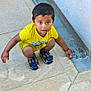 child, boy, yellow_clothing, sandals, water, curious, outdoor, pavement, flow, crouching, playing, concrete, young, person, cute, exploring, daylight, expression, small, fun