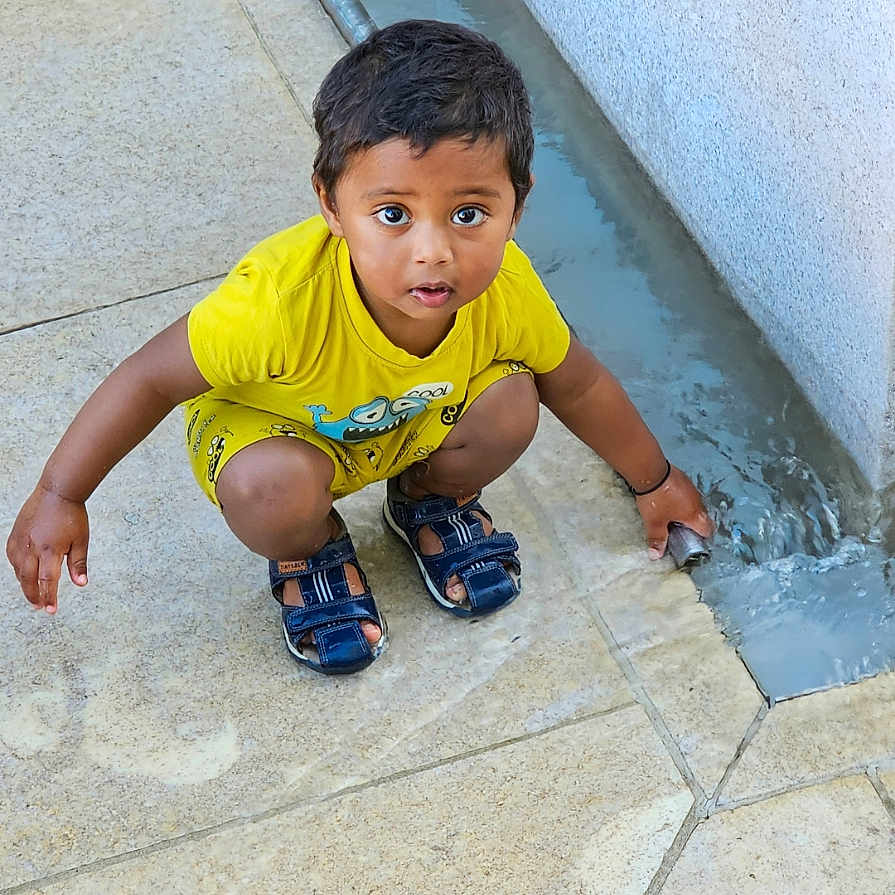 Riyansh a rejoint le concours — aidez-le/la à gagner de superbes lots ! boy, child, concrete, crouching, curious, cute, daylight, exploring, expression, flow, fun, outdoor, pavement, person, playing, sandals, small, water, yellow_clothing, young