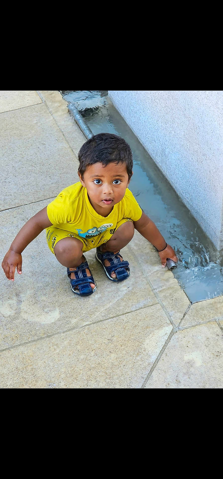 Riyansh a rejoint le concours — aidez-le/la à gagner de superbes lots ! child, boy, yellow_clothing, sandals, water, curious, outdoor, pavement, flow, crouching, playing, concrete, young, person, cute, exploring, daylight, expression, small, fun