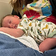 Owen is registered to the contest to win money with this photo: baby, sleeping, infant, dinosaur_onesie, clothing, jeans, tie_dye_shirt, alien_graphic, person, lap, resting, cozy, cute, indoor, portrait, child, peaceful, skin, hand, fabric