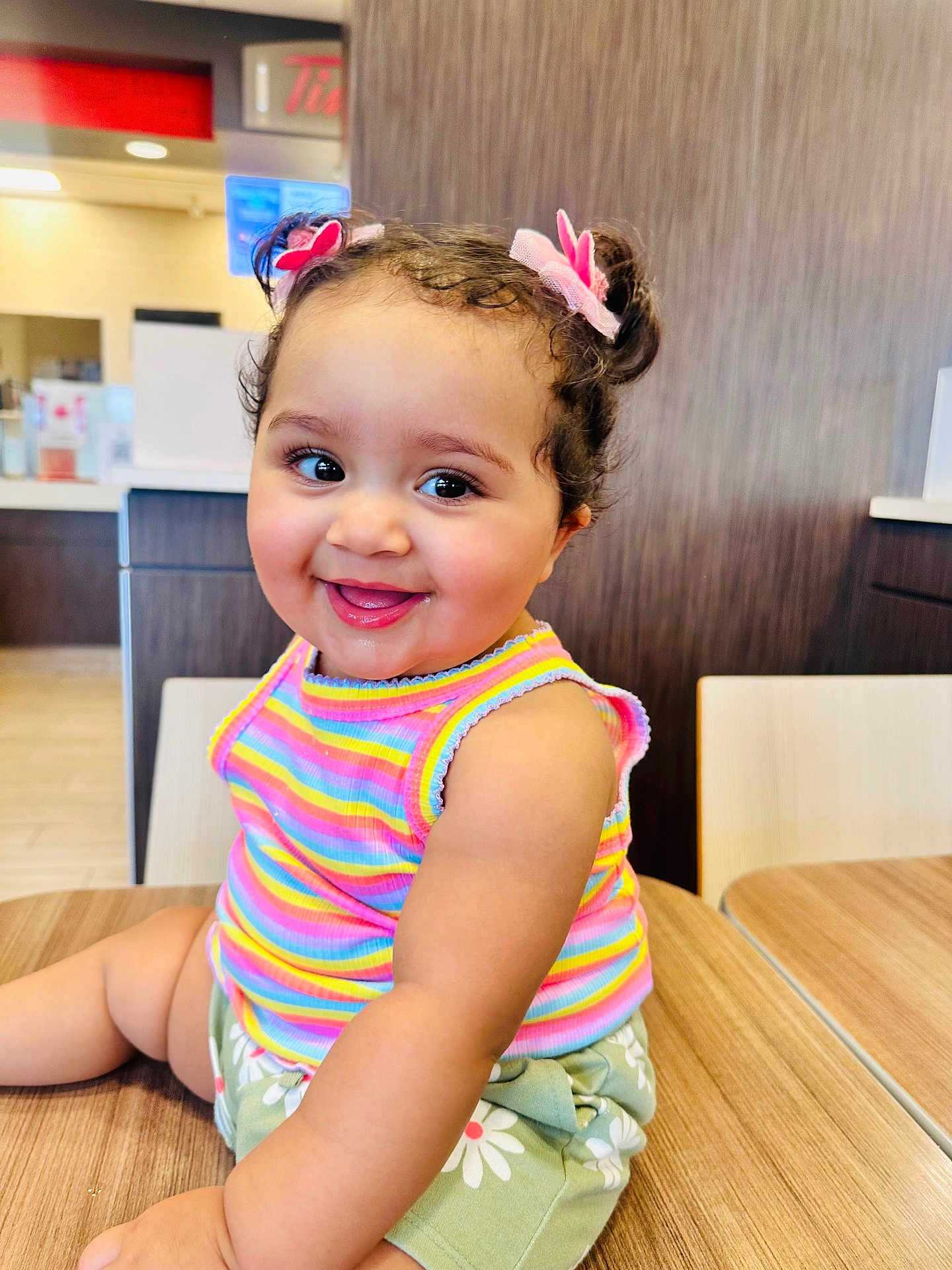 Fariba Binte Jamil joined the competition — help win amazing prizes! toddler, child, smile, happy, indoor, table, wooden_table, clothing, tank_top, shorts, hair_buns, hair_accessories, flower_clips, face, eyes, cheerful, cute, person, seated, daisies