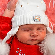 Paisley joined the competition — help win amazing prizes! baby, sleeping, white_hat, red_clothing, pom_poms, close_up, face, infant, soft_texture, cozy, peaceful, child, cute, portrait, headwear, newborn, resting, background, warm_colors, indoor