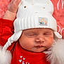 baby, sleeping, white_hat, red_clothing, pom_poms, close_up, face, infant, soft_texture, cozy, peaceful, child, cute, portrait, headwear, newborn, resting, background, warm_colors, indoor