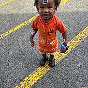 Christopher joined the competition — help win amazing prizes! child, boy, parking_lot, yellow_lines, orange_apron, stickers, curly_hair, black_crocs, shorts, orange_shirt, smiling, outdoor, standing, playful, face_stickers, casual_clothing, daylight, tarmac, young_child, happy