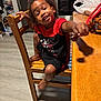 Christopher is registered to the contest to win money with this photo: child, curly_hair, smiling, lollipop, wooden_chair, kitchen, table, toy, plastic_bag, foot, floor, reaching_out, black_and_red_clothing, happy, indoor, appliance, cabinet, shadow, person, casual