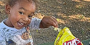Christopher joined the competition — help win amazing prizes! child, toddler, smiling, outdoor, park, picnic_table, snack, chips, lays, food, water_bottle, happy, tree, grass, daylight, casual_clothing, playful, young_child, person, nature