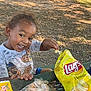 child, toddler, smiling, outdoor, park, picnic_table, snack, chips, lays, food, water_bottle, happy, tree, grass, daylight, casual_clothing, playful, young_child, person, nature