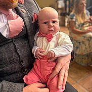 Jenson-James is registered to the contest to win money with this photo: baby, man, adult, person, holding, pink_bow_tie, pink_suspender, formal_wear, beard, pacifier, indoor, blurred_background, woman, seated, floor_tile, hand, shirt, vest, tie, face