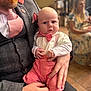 baby, man, adult, person, holding, pink_bow_tie, pink_suspender, formal_wear, beard, pacifier, indoor, blurred_background, woman, seated, floor_tile, hand, shirt, vest, tie, face