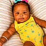 baby, child, infant, yellow_outfit, hair_bows, cushions, striped_pillow, relaxing, cute, smiling, close_up, indoors, skin, arm_jewelry, portrait, bright_colors, toddler, happy, plush, comfortable