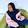 child, girl, black_hair, pink_jacket, black_shirt, green_chair, seated, indoor, casual_clothing, long_hair, portrait, relaxed, looking_away, young_person, modern_furniture, fashion, contemplative, comfortable, home, person
