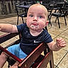 baby, child, high_chair, food, messy, blue_eyes, navy_shirt, light_blue_shorts, wooden_furniture, restaurant, tiled_floor, curious, tongue_out, indoor, person, seated, table, chair, casual, dining_area