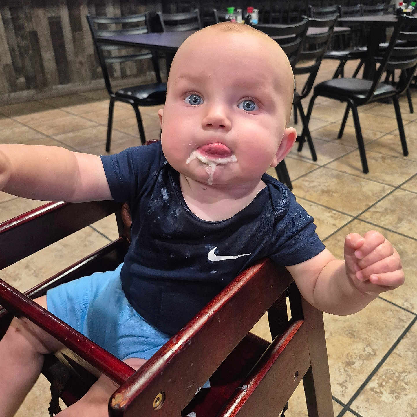 Virgil joined the competition — help win amazing prizes! baby, child, high_chair, food, messy, blue_eyes, navy_shirt, light_blue_shorts, wooden_furniture, restaurant, tiled_floor, curious, tongue_out, indoor, person, seated, table, chair, casual, dining_area