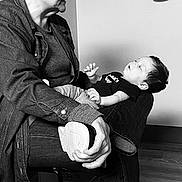 Robert Ray is registered to the contest to win money with this photo: baby, adult, woman, holding, sitting, lap, chair, cap, hat, denim_jacket, onesie, shoe, hand, indoor, wall, floor, black_and_white, portrait, candid, family