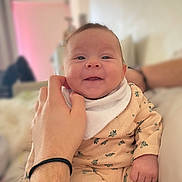 Luna participe au concours pour gagner de l'argent avec cette photo : adult_hand, baby, bed, bib, closeup, cozy, cute, face, floral_pattern, hair_tie, hand, happy, home_interior, infant, onesie, parent, pillow, portrait, smiling, wristband