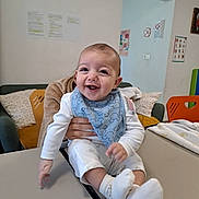 José a rejoint le concours — aidez-le/la à gagner de superbes lots ! baby, child, smiling, blue_bib, table, hand, indoor, couch, pillow, wall_poster, white_clothing, happy, sitting, person, sock, face, infant, support, room, furniture