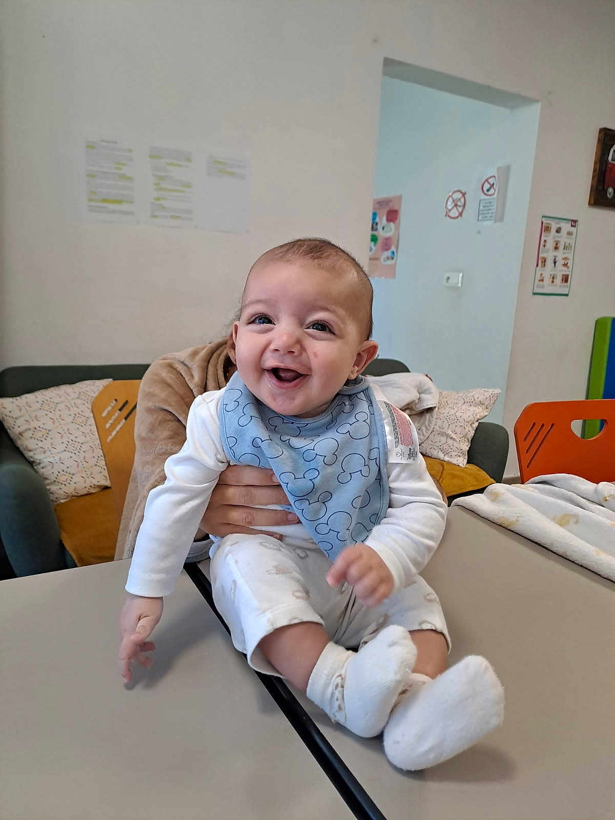 José a rejoint le concours — aidez-le/la à gagner de superbes lots ! baby, child, smiling, blue_bib, table, hand, indoor, couch, pillow, wall_poster, white_clothing, happy, sitting, person, sock, face, infant, support, room, furniture