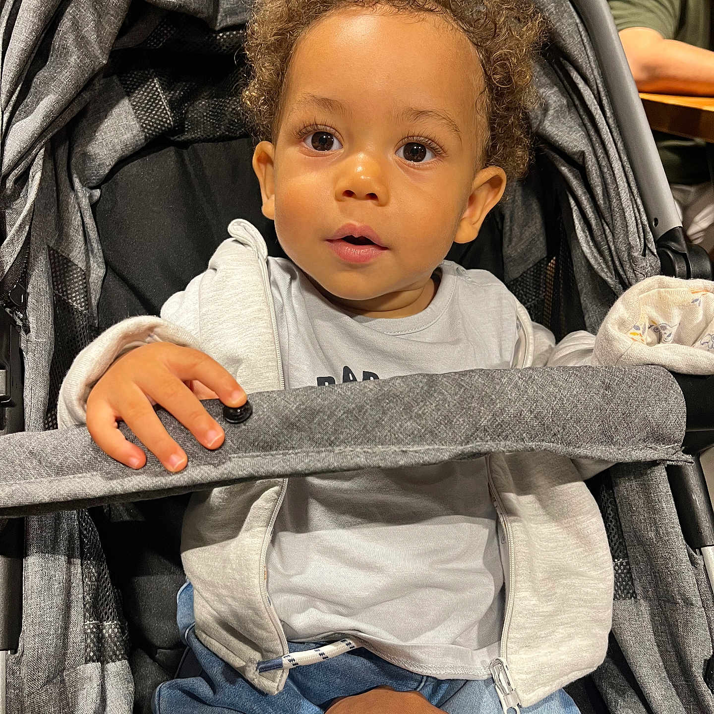 Maylann participe au concours pour gagner de l'argent avec cette photo : baby, blue_pants, casual_clothing, child, curiosity, curly_hair, cute, expression, face, gray_hoodie, hand, indoor, light, person, portrait, safety_bar, seated, stroller, support, toddler