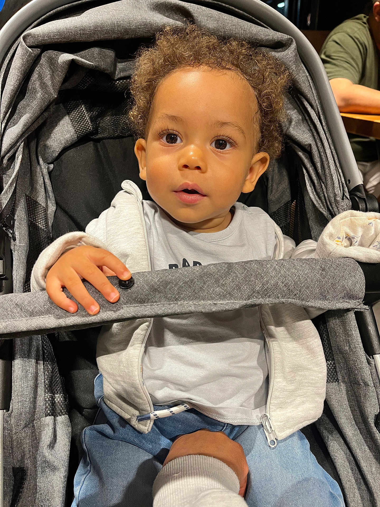 Maylann participe au concours pour gagner de l'argent avec cette photo : toddler, child, stroller, hand, curly_hair, gray_hoodie, blue_pants, safety_bar, indoor, person, seated, baby, expression, support, curiosity, casual_clothing, light, face, portrait, cute