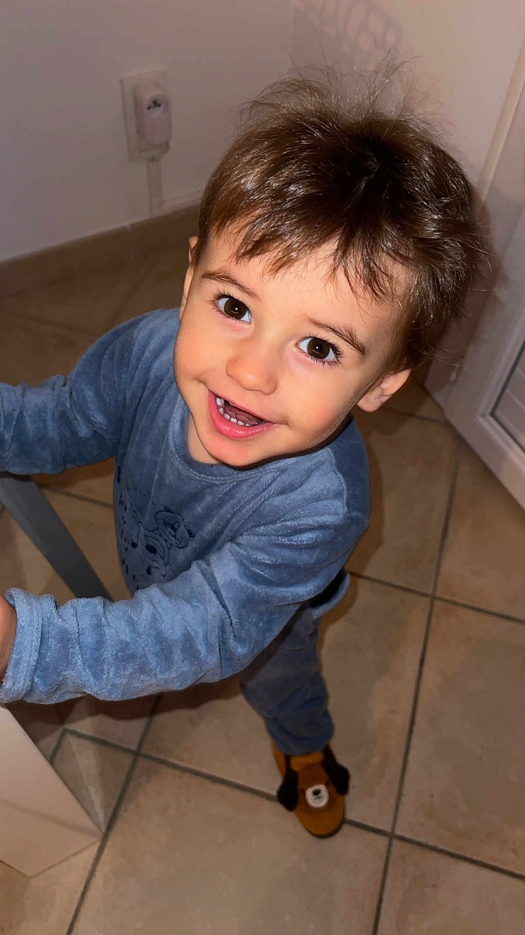 Alessio participe au concours pour gagner de l'argent avec cette photo : brown_hair, child, cute, door, electric_outlet, floor, footwear, happy, home, indoor, long_sleeves, looking_up, pajamas, person, slippers, smiling, standing, tile, toddler, young_child