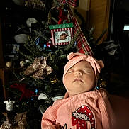 Amelia is registered to the contest to win money with this photo: baby, sleeping, pink_clothing, headwear, christmas_tree, ornaments, holiday_decor, indoor, festive, portrait, child, soft_lighting, celebration, holiday_season, infant, cute, cozy, family, seasonal, decorations