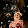 baby, sleeping, pink_clothing, headwear, christmas_tree, ornaments, holiday_decor, indoor, festive, portrait, child, soft_lighting, celebration, holiday_season, infant, cute, cozy, family, seasonal, decorations