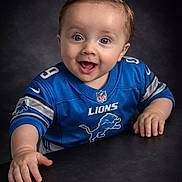 Frankie is registered to the contest to win money with this photo: baby, child, smiling, blue_jersey, detroit_lions, sportswear, portrait, happy, face, hands, indoor, studio_lighting, cute, young_child, person, expression, short_hair, looking_at_camera, sitting, background
