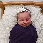 Harper is registered to the contest to win money with this photo: newborn, baby, swaddled, purple_wrap, white_flower_headband, wooden_bed, fluffy_cushion, sleeping, smiling, infant, cute, portrait, soft_lighting, indoors, cozy, peaceful, headband, child, person, babywearing