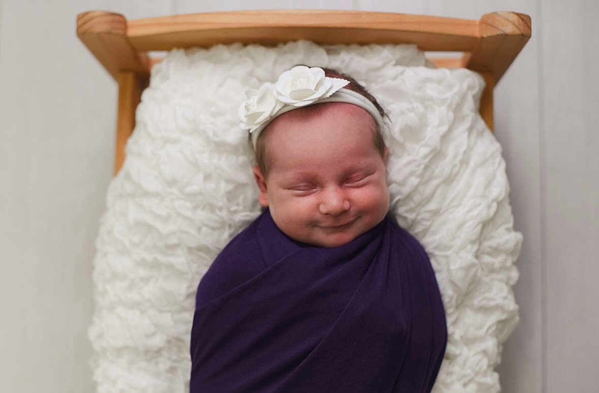 Harper is registered to the contest to win money with this photo: newborn, baby, swaddled, purple_wrap, white_flower_headband, wooden_bed, fluffy_cushion, sleeping, smiling, infant, cute, portrait, soft_lighting, indoors, cozy, peaceful, headband, child, person, babywearing