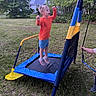 Oliver is registered to the contest to win money with this photo: child, trampoline, jumping, outdoor, grass, trees, house, person, rope, playground, blue, yellow, orange_shirt, shorts, sneakers, backyard, nature, fun, active, daytime