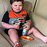 child, boy, couch, snack, sippy_cup, orange_shirt, black_shorts, indoor, smile, face, hands, legs, barefoot, beige, furniture, casual, relaxed, portrait, home, cute