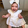 baby, bow, carpet, child, chubby_arms, cute, face, fashion, footwear, happy, headband, indoor, infant, person, portrait, sitting, smiling, soft_lighting, white_dress, young_child