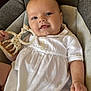 baby, infant, smiling, white_dress, giraffe_toy, teether, cushioned_seat, indoor, child, cute, relaxed, happy, portrait, person, furniture, skin, hand, head, eyes, mouth