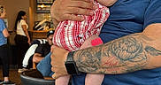 Athena is registered to the contest to win money with this photo: baby, blue, cafe, casual, child, clothing, curious, expression, gray, hat, headwear, holding, indoor, man, people, person, portrait, red, tattoo, watch