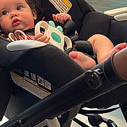 Athena joined the competition — help win amazing prizes! baby, car_seat, toy, indoor, child, curly_hair, black_clothing, stroller, floor, window, chair, light, hand, foot, face, person, seatbelt, warning_label, modern, curious