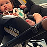 baby, car_seat, toy, indoor, child, curly_hair, black_clothing, stroller, floor, window, chair, light, hand, foot, face, person, seatbelt, warning_label, modern, curious
