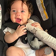 Athena joined the competition — help win amazing prizes! baby, child, car_seat, smiling, happy, curly_hair, toy, infant, seatbelt, white_clothing, cute, safety, closeup, face, skin, small_hands, indoors, car, child_safety, expression