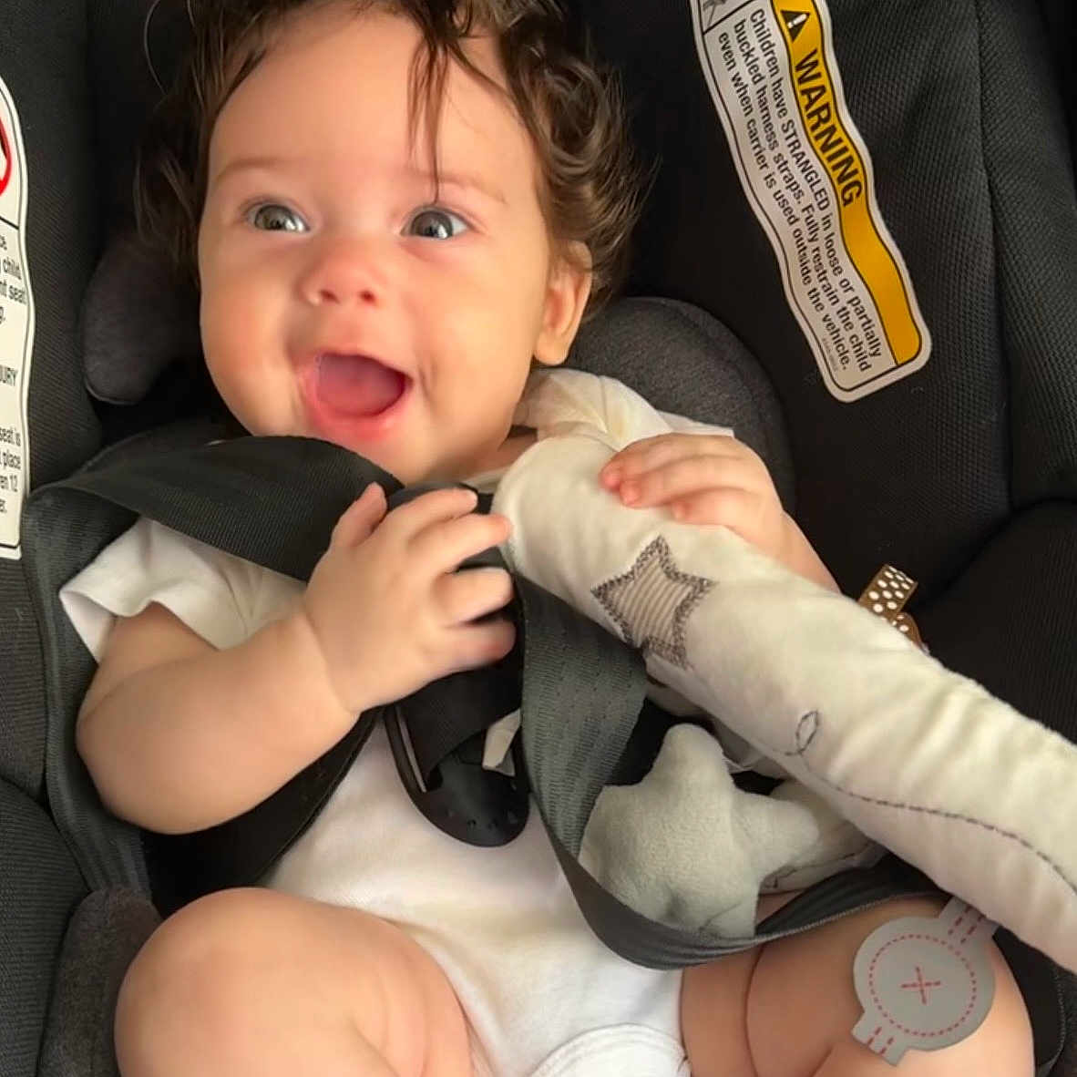Athena joined the competition — help win amazing prizes! baby, child, car_seat, smiling, happy, curly_hair, toy, infant, seatbelt, white_clothing, cute, safety, closeup, face, skin, small_hands, indoors, car, child_safety, expression