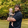Dallas Kash is registered to the contest to win money with this photo: adult_male, baby, child, smiling, beard, black_shirt, plaid_shirt, outdoor, grass, trees, nature, portrait, holding, happy, family, casual_clothing, daylight, person, father_and_son, barefoot