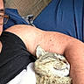 arm, bed_sheet, black_tank_top, blue_bed_sheet, cat, close_up, comfort, content, cozy, cuddle, fur, glasses, indoor, person, pet, relaxation, resting, sleeping, tabby_cat, wooden_floor