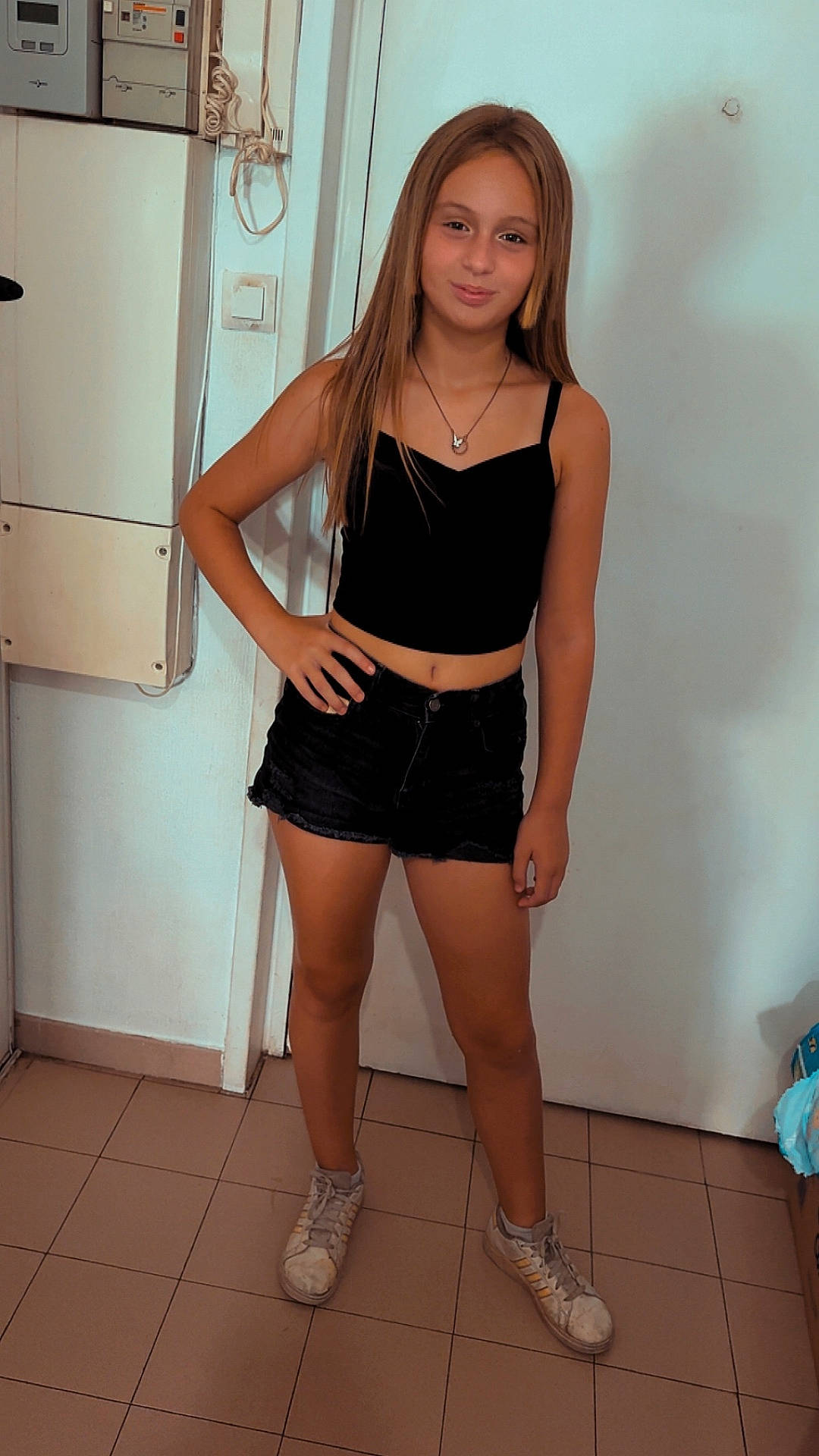 black_shorts, black_top, casual_clothing, child, door, footwear, girl, hand_on_hip, indoor, long_hair, necklace, person, portrait, pose, smile, sneakers, standing, tile_floor, wall, young_person