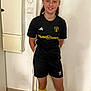 Serena a rejoint le concours — aidez-le/la à gagner de superbes lots ! child, soccer, sports_uniform, yellow_socks, black_shorts, black_shirt, indoor, standing, smiling, young_athlete, person, tile_floor, wall, door, short_hair, casual_pose, team_sport, athletic, ready, confident