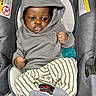baby, car_seat, casual_wear, child, clenched_fist, cute, dark_skin, expression, gray_clothing, hoodie, indoor, infant, pacifier, portrait, seatbelt, seated, socks, striped_pants, warning_label, young_child