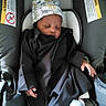 Saadiq is registered to the contest to win money with this photo: baby, infant, sleeping, car_seat, hat, clothing, black_outfit, dinosaur_pattern, warning_label, seatbelt, child, person, indoors, cute, peaceful, small_hand, soft_fabric, comfort, resting, newborn