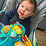 baby, baby_seat, blue_eyes, blue_tray, casual_clothing, child, child_in_seat, closeup, colorful_toys, curious, gray_seat, happy_baby, indoors, plastic_toys, playtime, seat_belt, smiling, toy, wooden_floor, young_child
